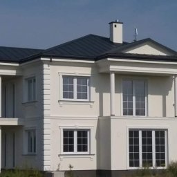 DMBudownictwo - Dwupiętrowy dom z kolumnami i ciemnym dachem, wykończony w jasnej kolorystyce, z widocznym kominem i anteną na dachu, otoczony zielenią.