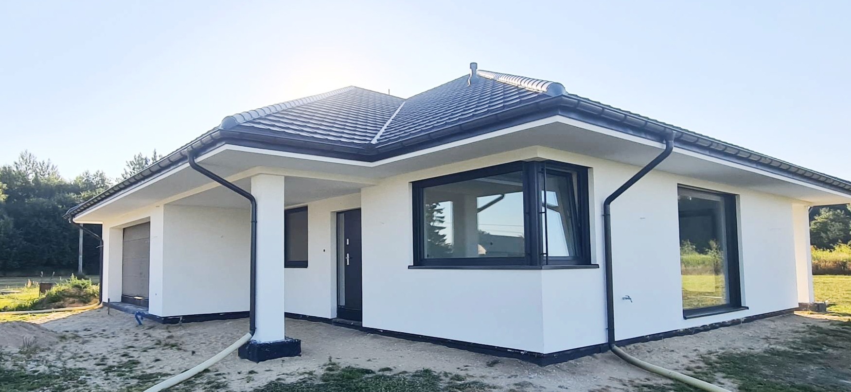 Nowoczesny, parterowy dom z ciemną dachówką i grafitową stolarką okienną, świeżo otynkowany na biało, z widocznymi rurami spustowymi i fragmentem terenu przed budynkiem.