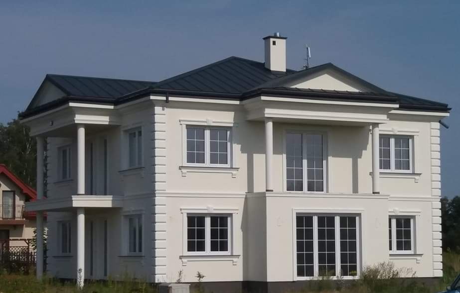 Dwupiętrowy dom z kolumnami i ciemnym dachem, wykończony w jasnej kolorystyce, z widocznym kominem i anteną na dachu, otoczony zielenią.