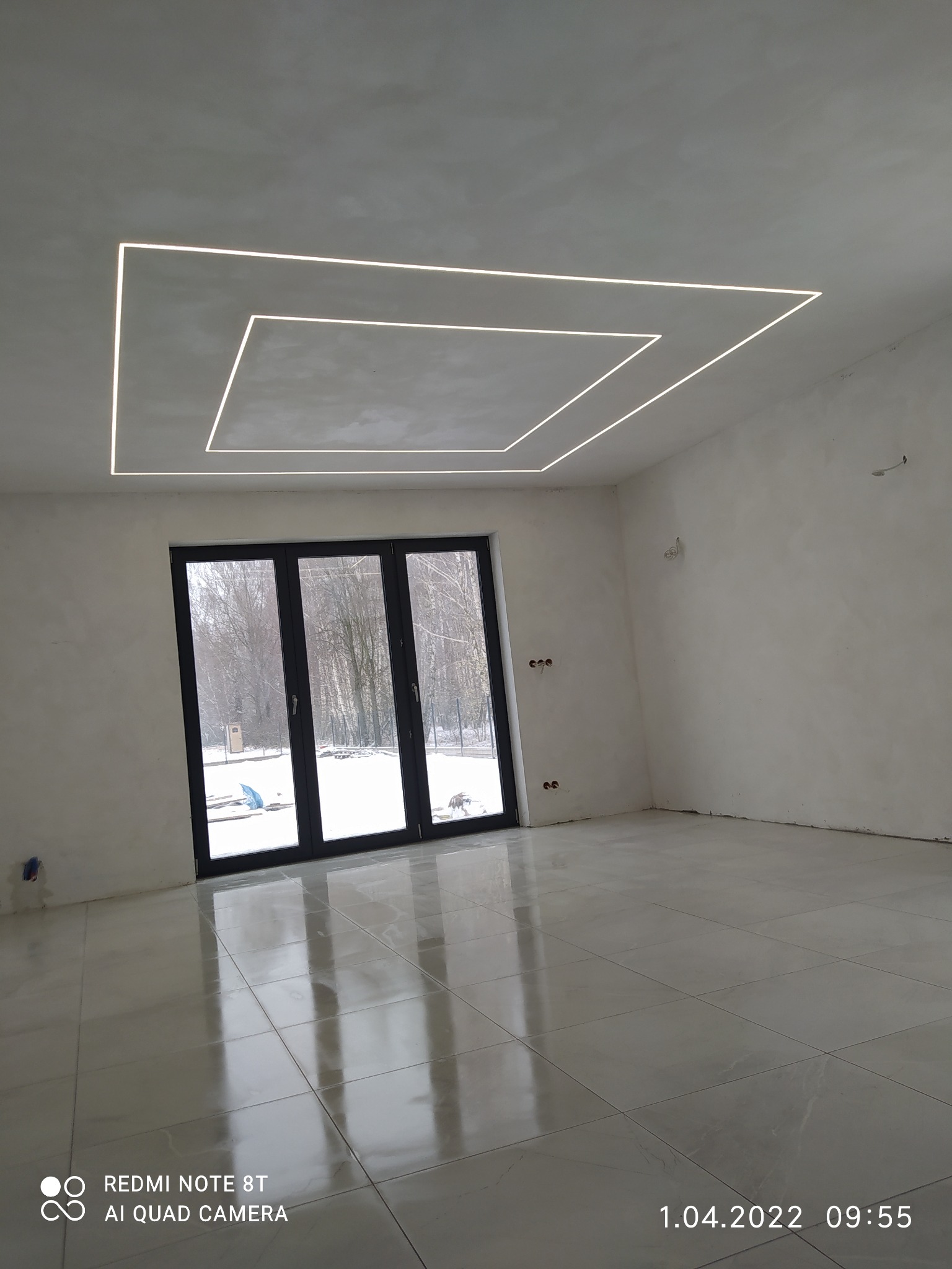 Pusty pokój z błyszczącą, jasną podłogą z płytek i nowoczesnym, kwadratowym oświetleniem LED w suficie, z widokiem na zimowy krajobraz przez duże okno balkonowe.