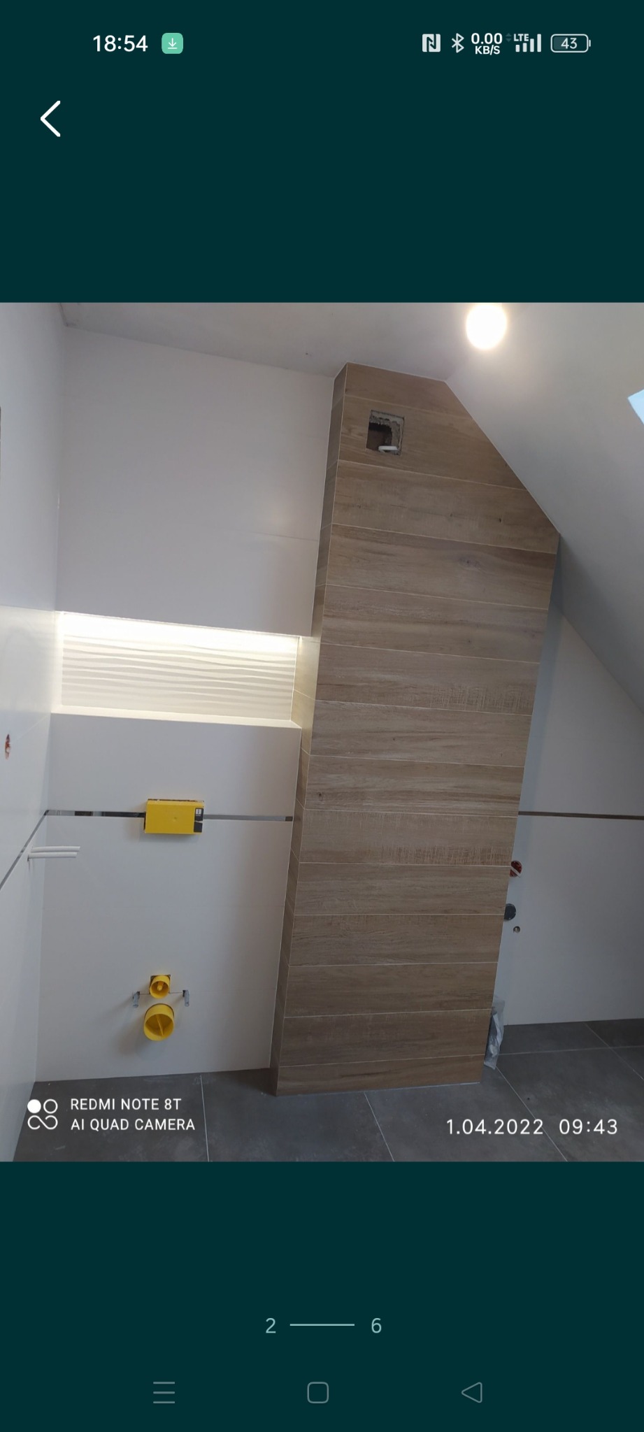 Ściana w łazience w trakcie prac wykończeniowych, widoczne płytki imitujące drewno na skośnej zabudowie, instalacja podtynkowa WC z żółtymi elementami, białe ściany i sufit ze światłem led.