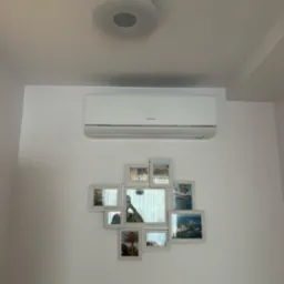 Biała klimatyzacja marki Hisense zamontowana na białej ścianie, poniżej kolaż z ramek z widokiem na morze i palmy, okrągła lampa sufitowa w tle.