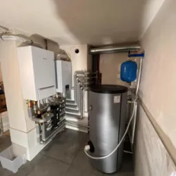 Zestaw dwóch wiszących kotłów grzewczych marki Samsung i Vaillant, połączonych z szarym zasobnikiem ciepłej wody i niebieskim naczyniem wzbiorczym, w pomieszczeniu z szarą podłogą i widocznymi...