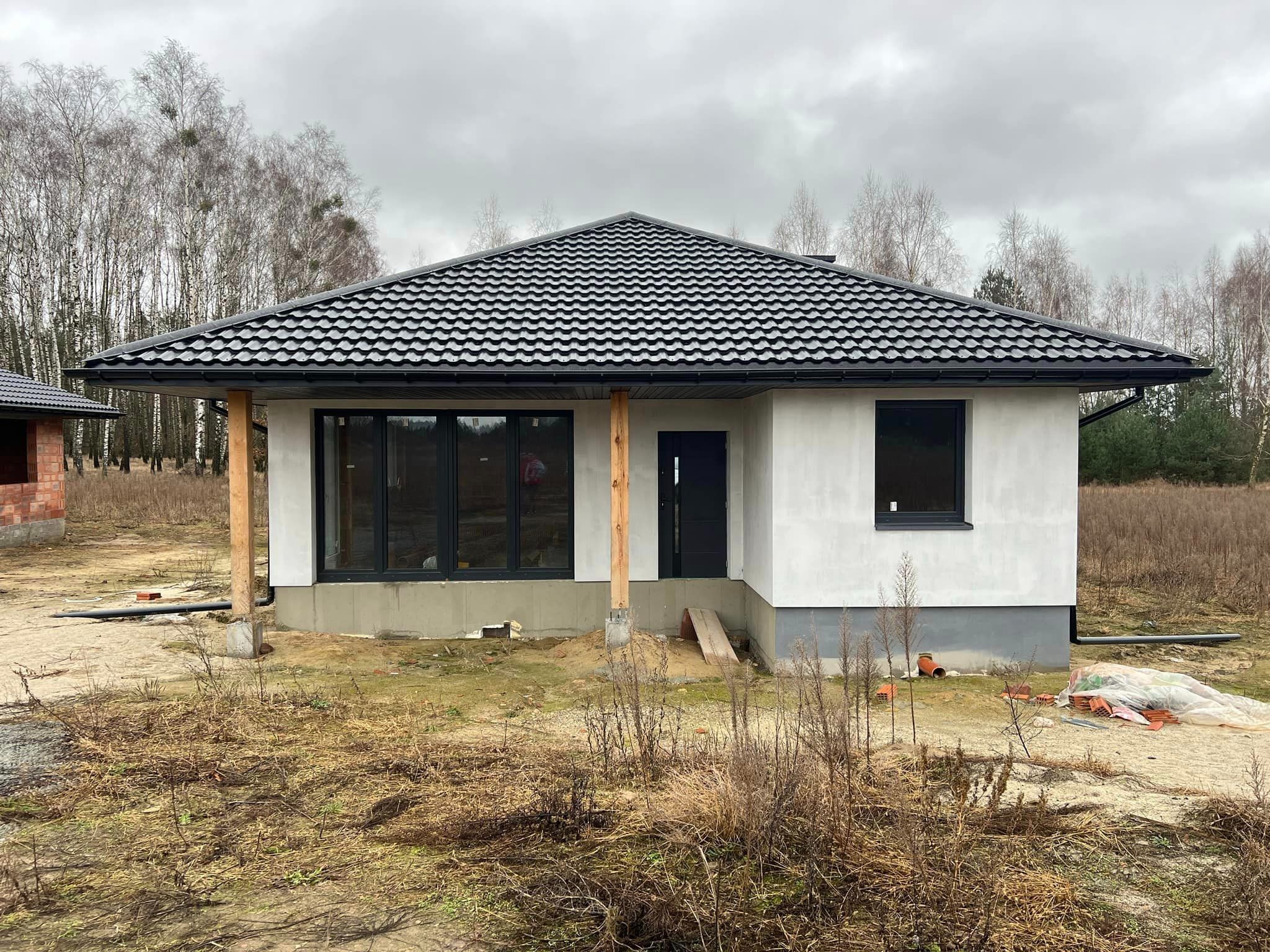 Surowy, parterowy dom murowany z czarną dachówką, surowymi drewnianymi słupami i szarym fundamentem, w otoczeniu jesiennego krajobrazu z widocznymi rurami i materiałami budowlanymi.