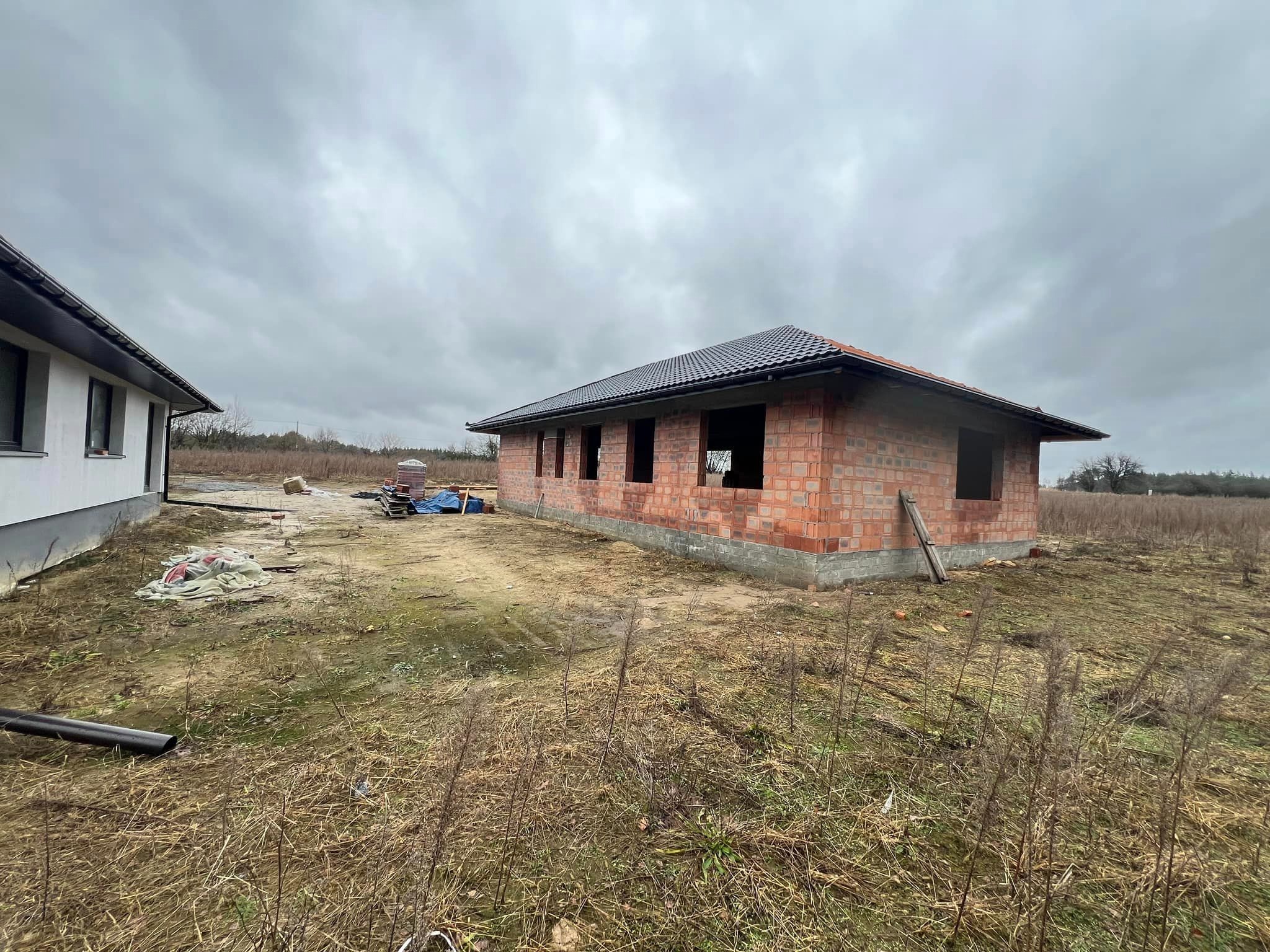 Surowy stan domu z czerwonej cegły z częściowo ukończonym dachem na tle pochmurnego nieba, widoczne fundamenty i sąsiedni budynek.