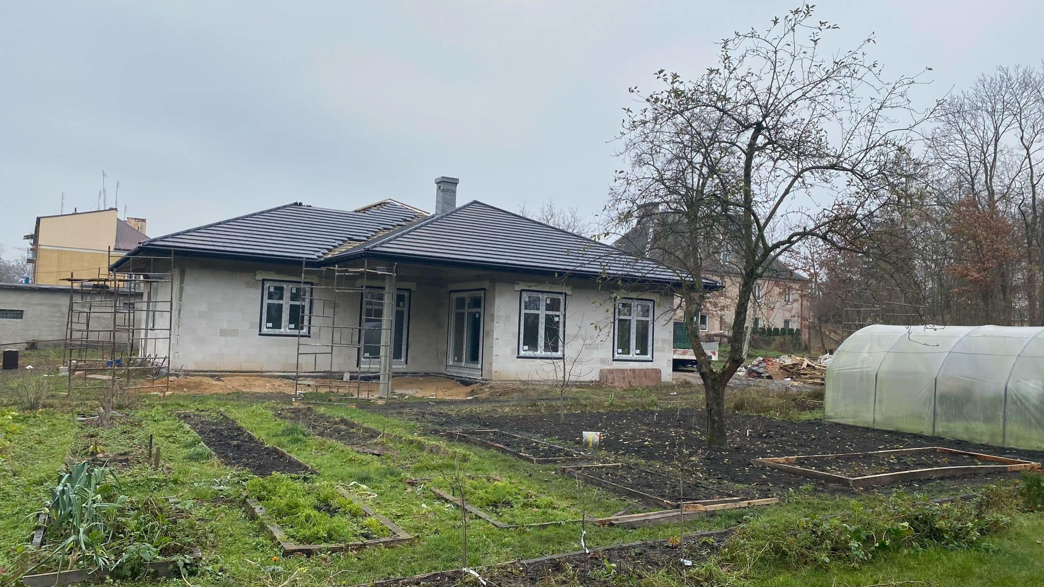 Surowy stan domu z bloczków z czarną dachówką, widoczne rusztowania, ogród warzywny w pierwszym planie i szklarnia w tle.