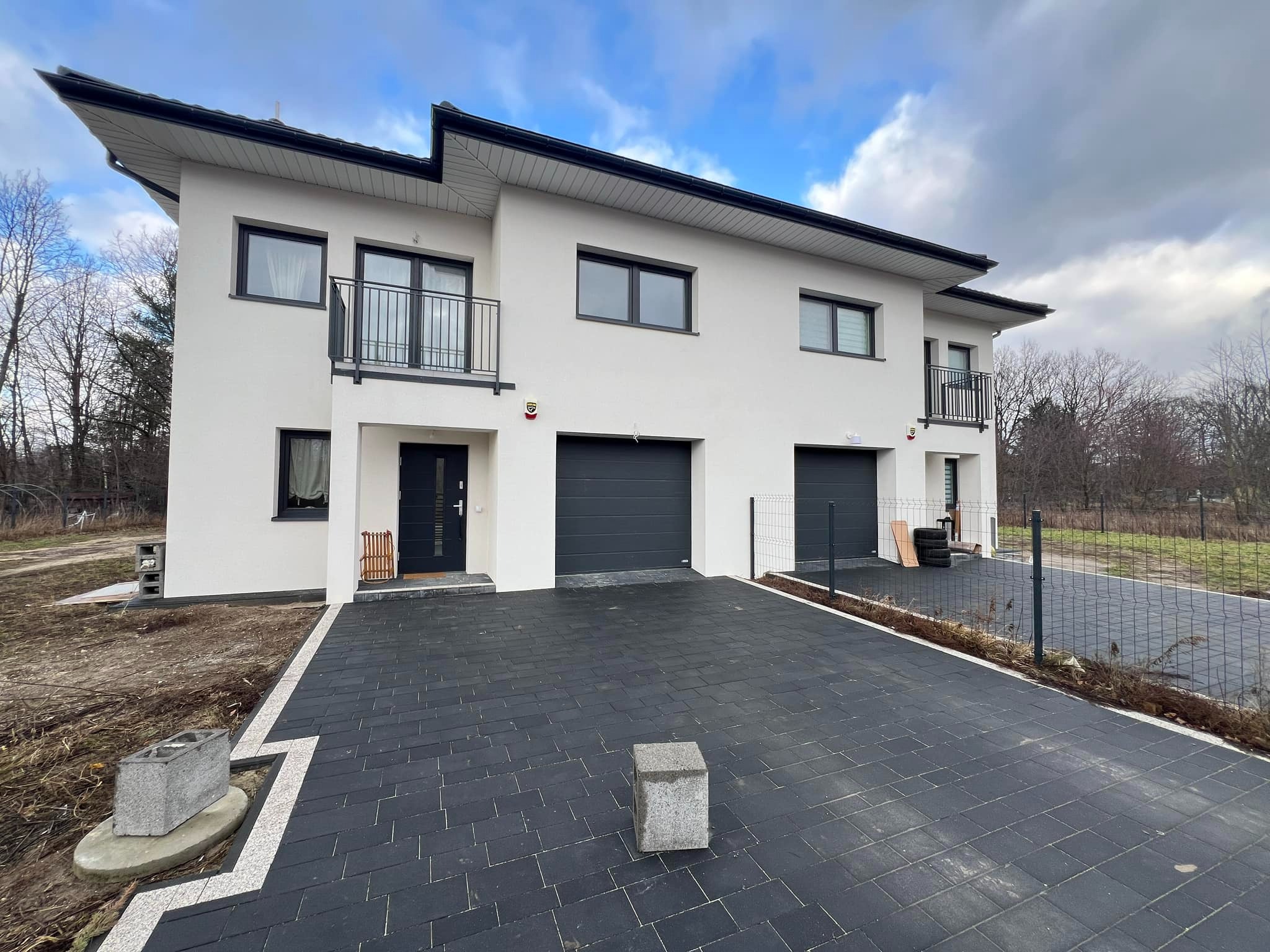 Nowoczesny, dwukondygnacyjny dom bliźniak z białymi ścianami, czarnymi oknami i bramami garażowymi, wykończony czarną dachówką, z utwardzonym podjazdem z kostki brukowej i minimalistycznymi balkonami.