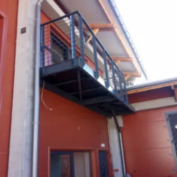 Metalowa platforma balkonowa z minimalistyczną balustradą z cienkich prętów, zamontowana na elewacji budynku o ceglastym kolorze, widok z dołu.