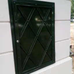 Zielona, metalowa klapka rewizyjna z ozdobnym kratowaniem, zamontowana w białej, murowanej konstrukcji na zewnątrz budynku.