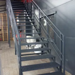 Zewnętrzne metalowe schody z ażurowymi stopniami i poręczami prowadzące w górę, przylegające do szarej hali z czerwonym akcentem, widoczne palety w tle.