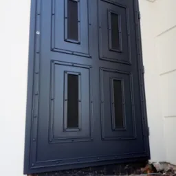 Ciemnoszare, metalowe drzwi zewnętrzne z czterema prostokątnymi, ażurowymi panelami, mocowane na zawiasach do białej ściany budynku. Widoczne ślady prac budowlanych u podstawy.