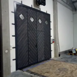 Dwuskrzydłowa brama garażowa z ciemnego drewna ułożonego w jodełkę, z czterema romboidalnymi okienkami, zamontowana w hali przemysłowej z widocznymi betonowymi słupami i kratką ściekową w posadzce.