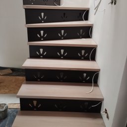 BSL Welding Art - Schody z jasnego drewna z czarnymi podstopnicami z wyciętymi wzorami, widoczne kable instalacyjne przy ścianie.