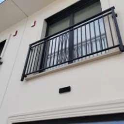 Elegancki balkon z czarną, metalową balustradą o dekoracyjnym wzorze, zamontowaną na jasnej elewacji budynku. Widoczne detale architektoniczne i okna.