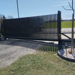 Nowoczesna, przesuwna brama wjazdowa z pionowych, metalowych profili, rzucająca wyraźny cień na podjazd. Za bramą widoczne zielone pole i ogrodzenie z siatki.