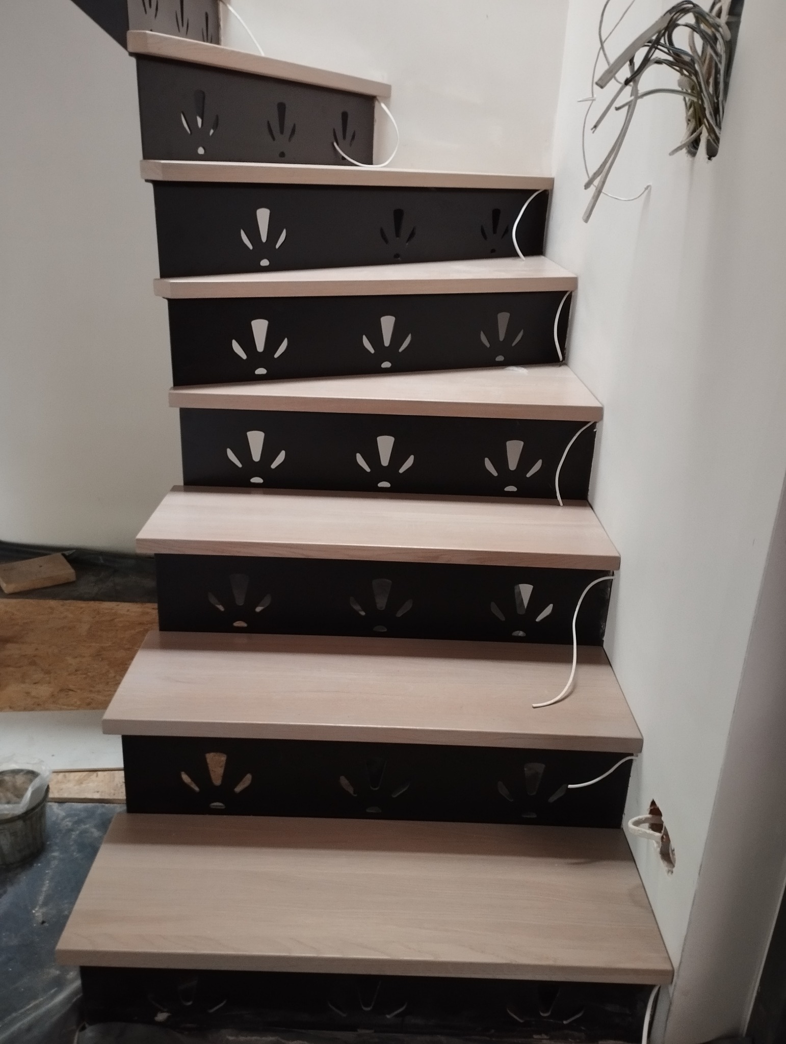 Schody z jasnego drewna z czarnymi podstopnicami z wyciętymi wzorami, widoczne kable instalacyjne przy ścianie.