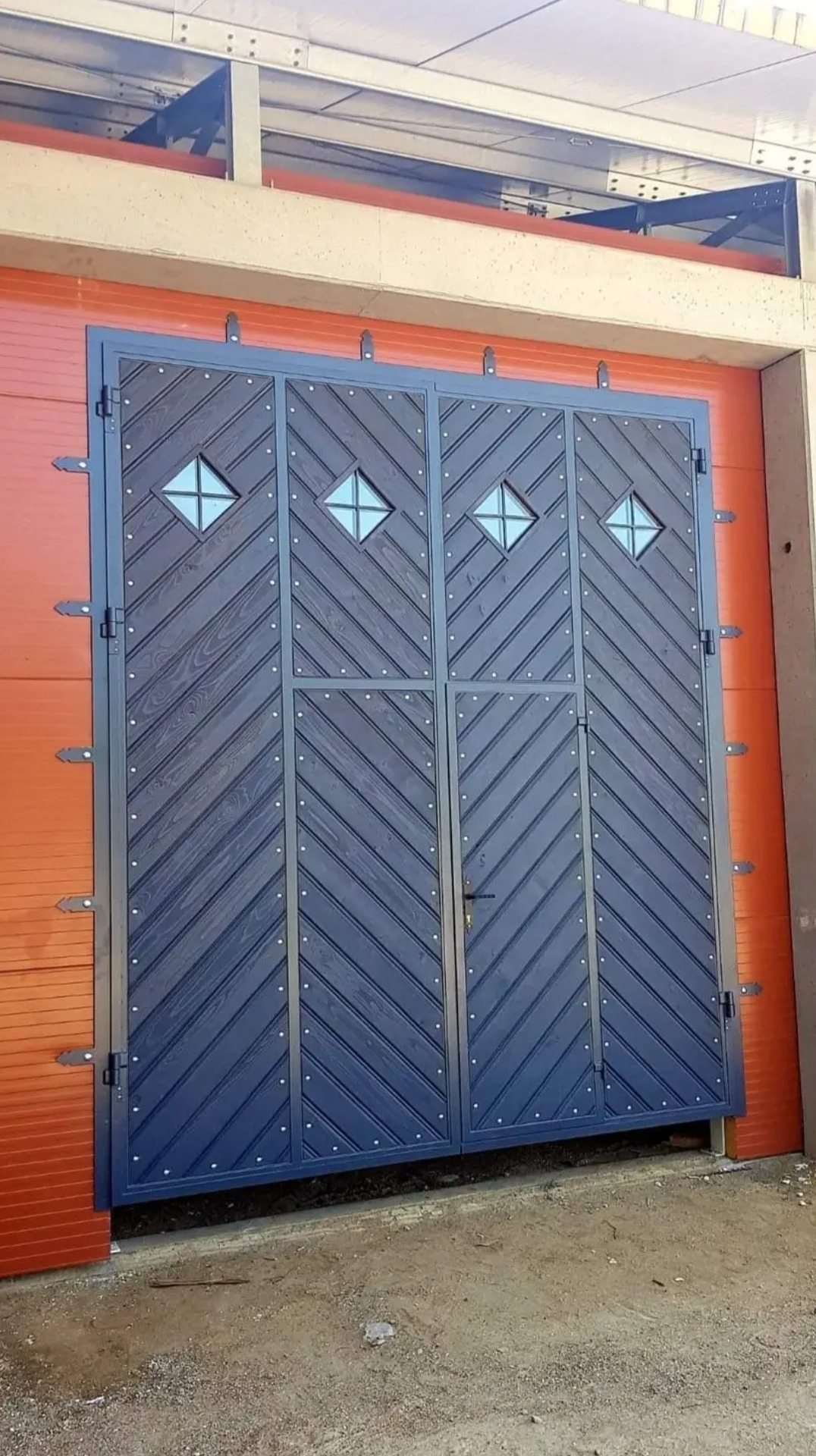 Dwuskrzydłowa brama wjazdowa z ciemnego drewna z metalową ramą i czterema kwadratowymi okienkami z szybami, zamontowana na pomarańczowej ścianie budynku.