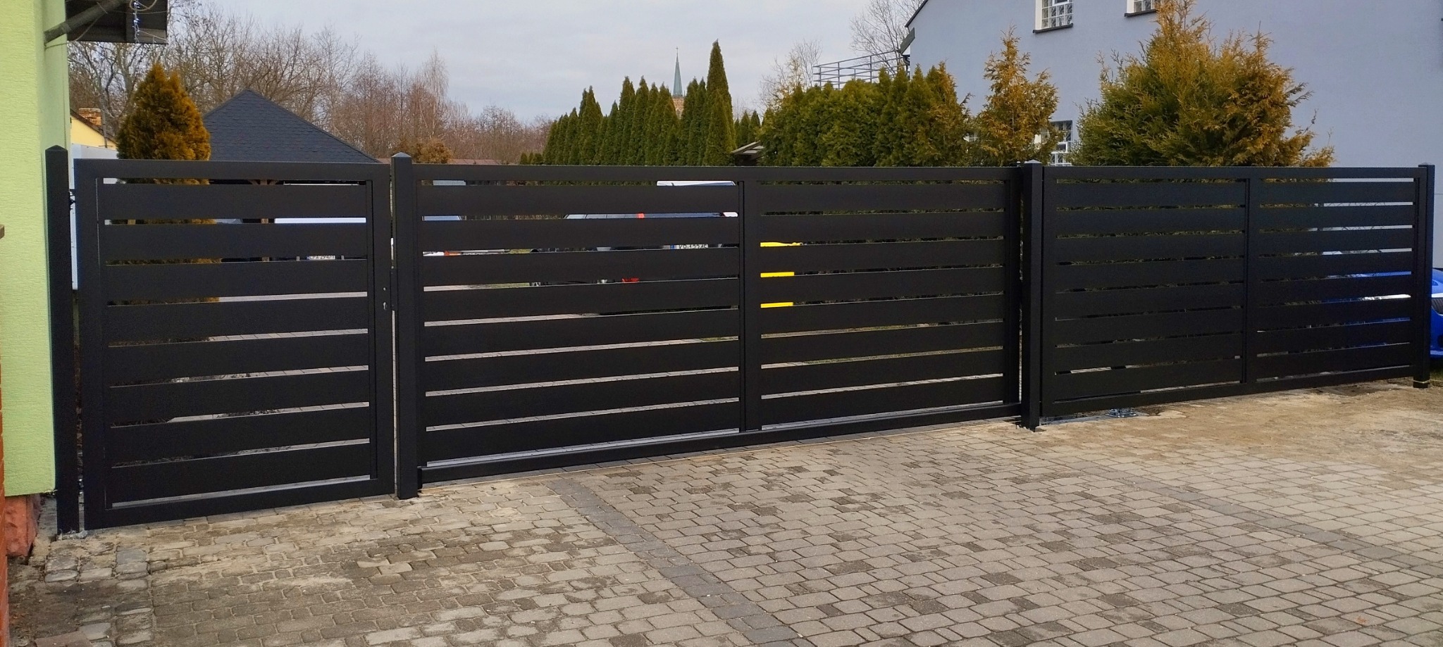Nowoczesna, przesuwna brama wjazdowa z poziomych, czarnych profili metalowych na tle brukowanej nawierzchni podjazdu i budynku mieszkalnego.