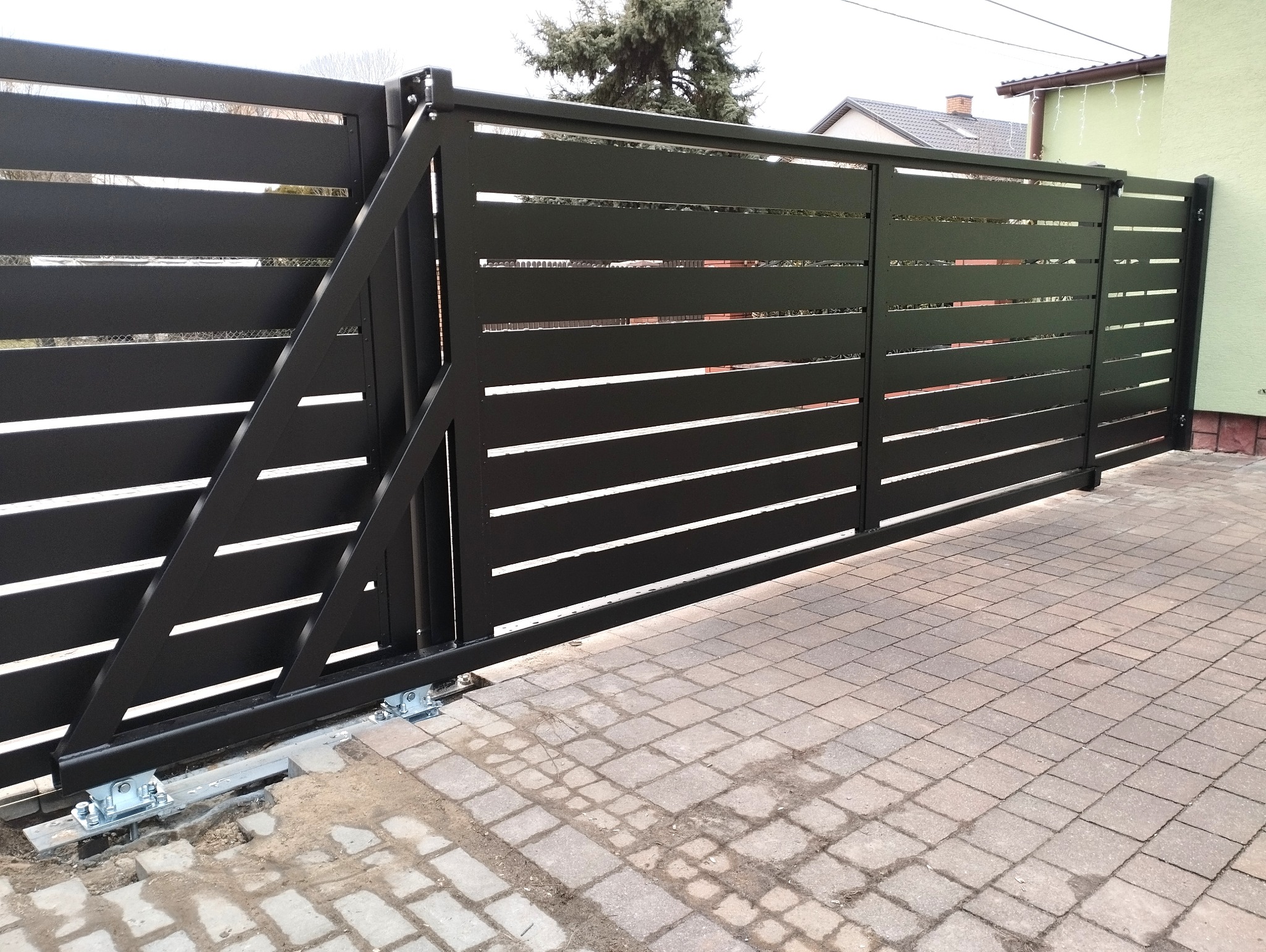 Czarna, przesuwna brama wjazdowa z poziomymi szczelinami, widoczne mechanizmy jezdne i kostka brukowa na podjeździe.