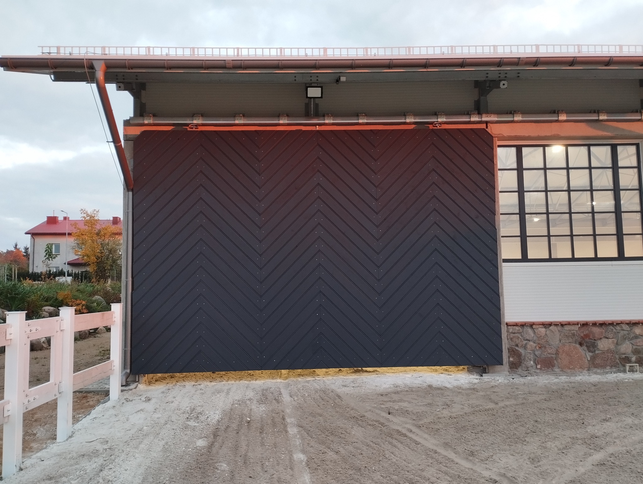 Nowoczesna, duża, przesuwna brama garażowa z czarnego metalu, z ukośnym wzorem desek, zamontowana w budynku z dużym oknem i kamienną podmurówką, widoczna z zewnątrz, z elementami otoczenia.