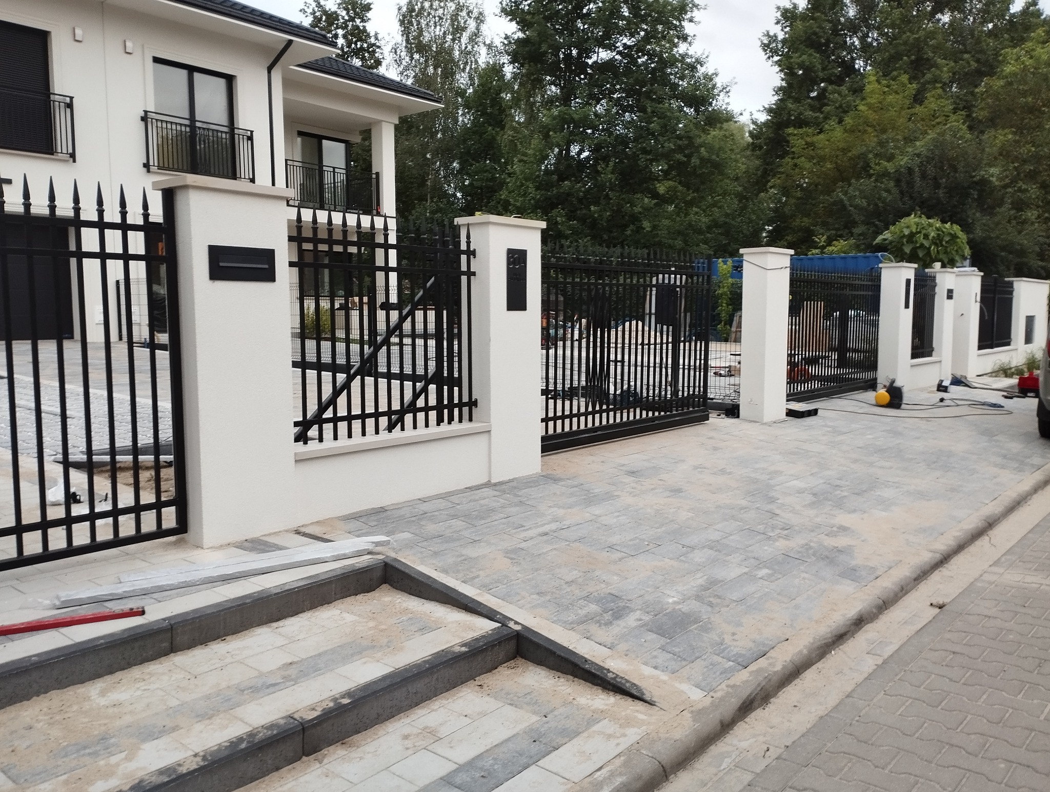 Nowoczesne ogrodzenie z czarnymi, metalowymi przęsłami i białymi słupkami. Widoczne schody z kostki brukowej prowadzące do bramy wjazdowej. Elementy montażowe na schodach.