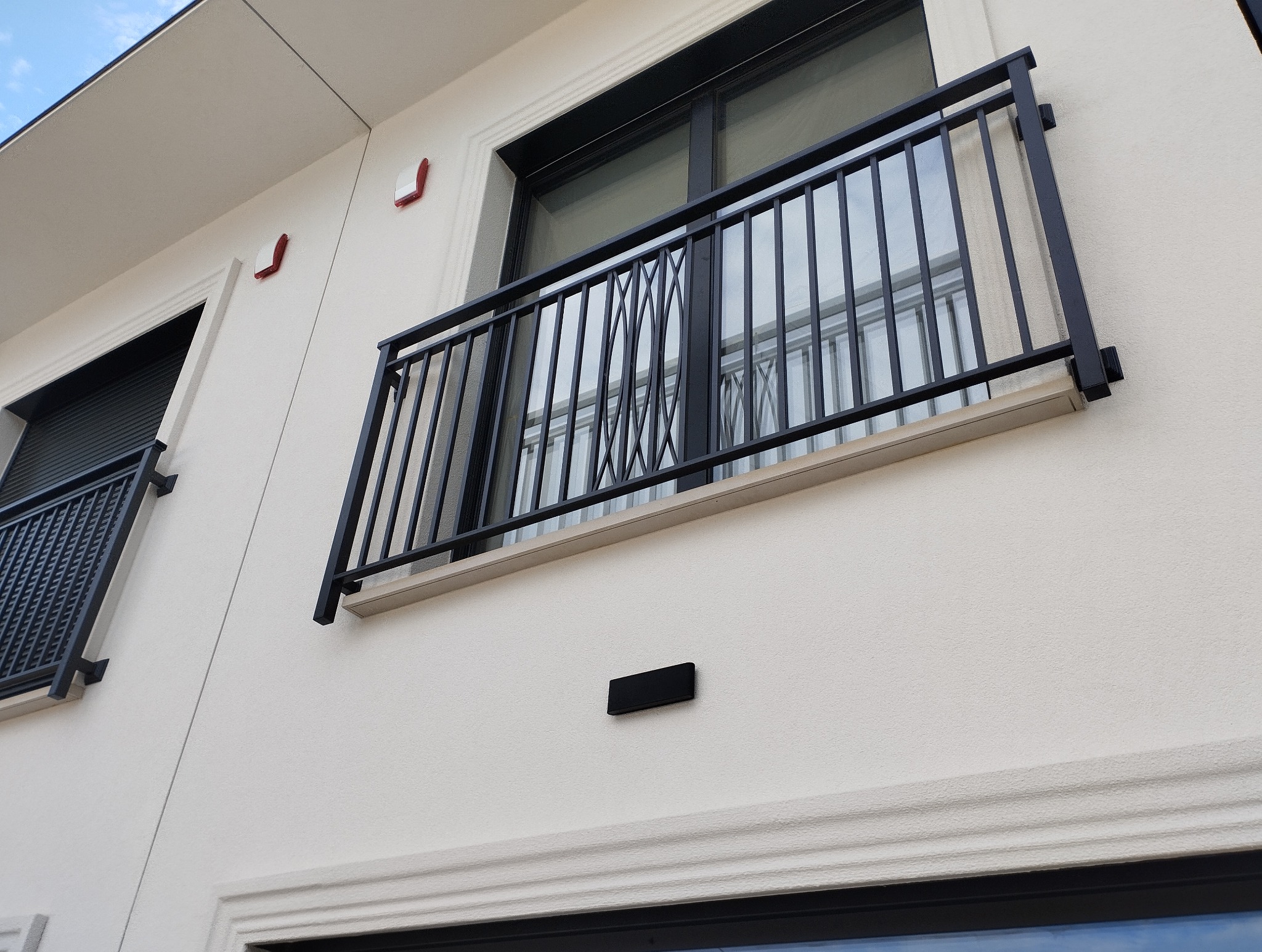 Elegancki balkon z czarną, metalową balustradą o dekoracyjnym wzorze, zamontowaną na jasnej elewacji budynku. Widoczne detale architektoniczne i okna.
