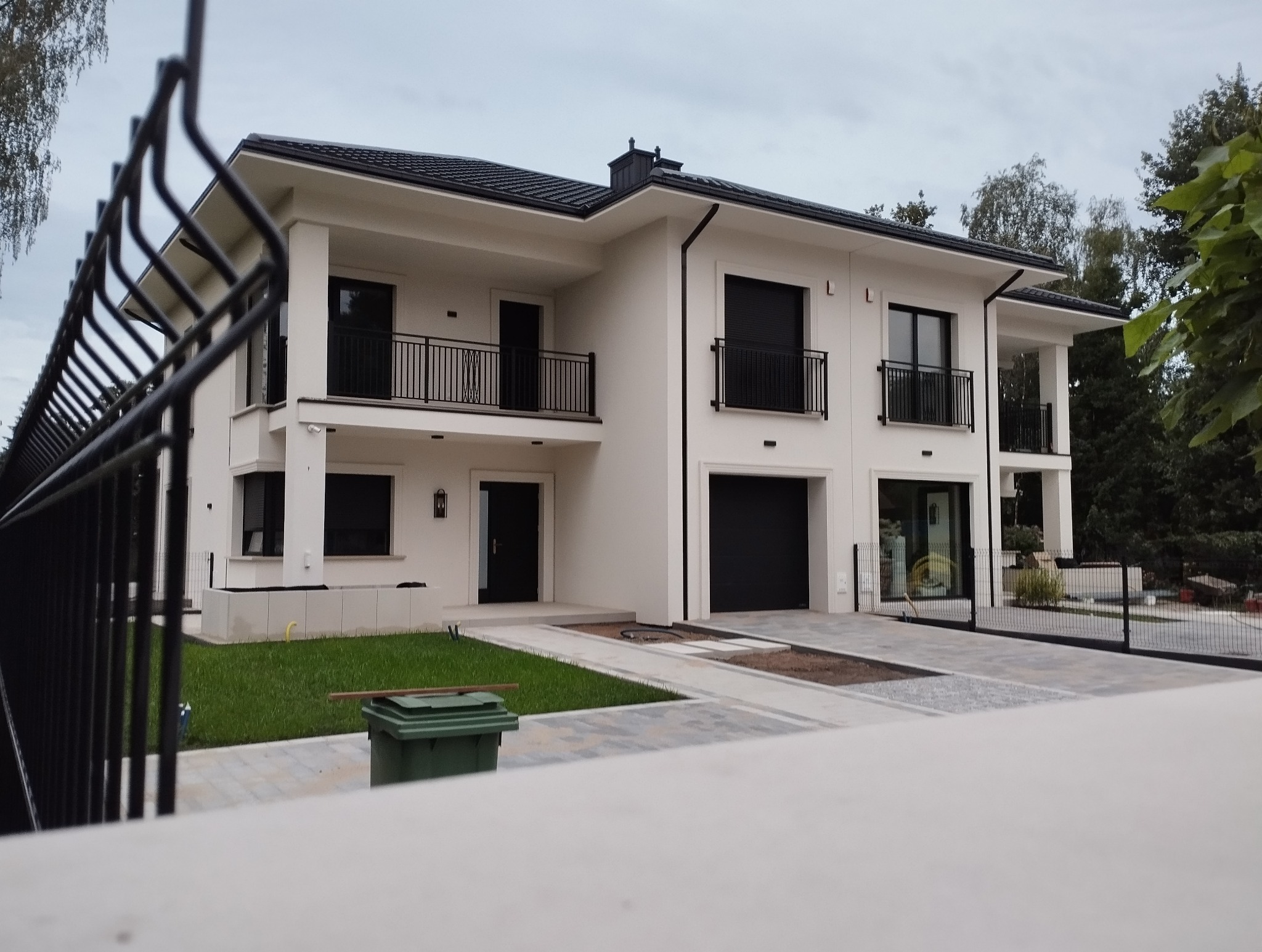Nowoczesny, dwukondygnacyjny dom z czarnymi detalami: balustrady balkonowe, brama garażowa i stolarka okienna. Widoczny fragment ogrodzenia panelowego.