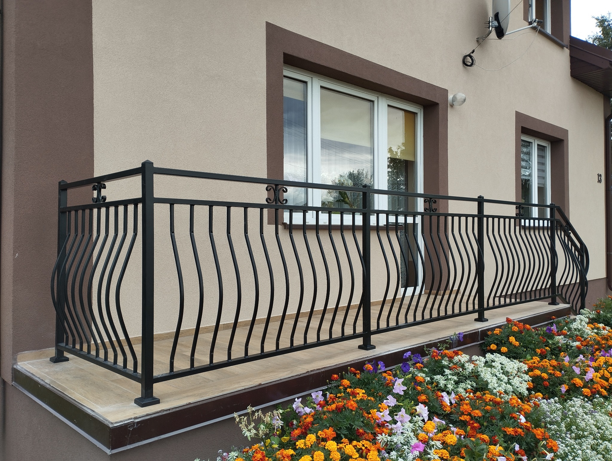 Eleganckie, czarne, metalowe balustrady balkonowe z dekoracyjnymi, falowanymi prętami, zdobiące balkon domu z beżową elewacją i kolorowym kwietnikiem.
