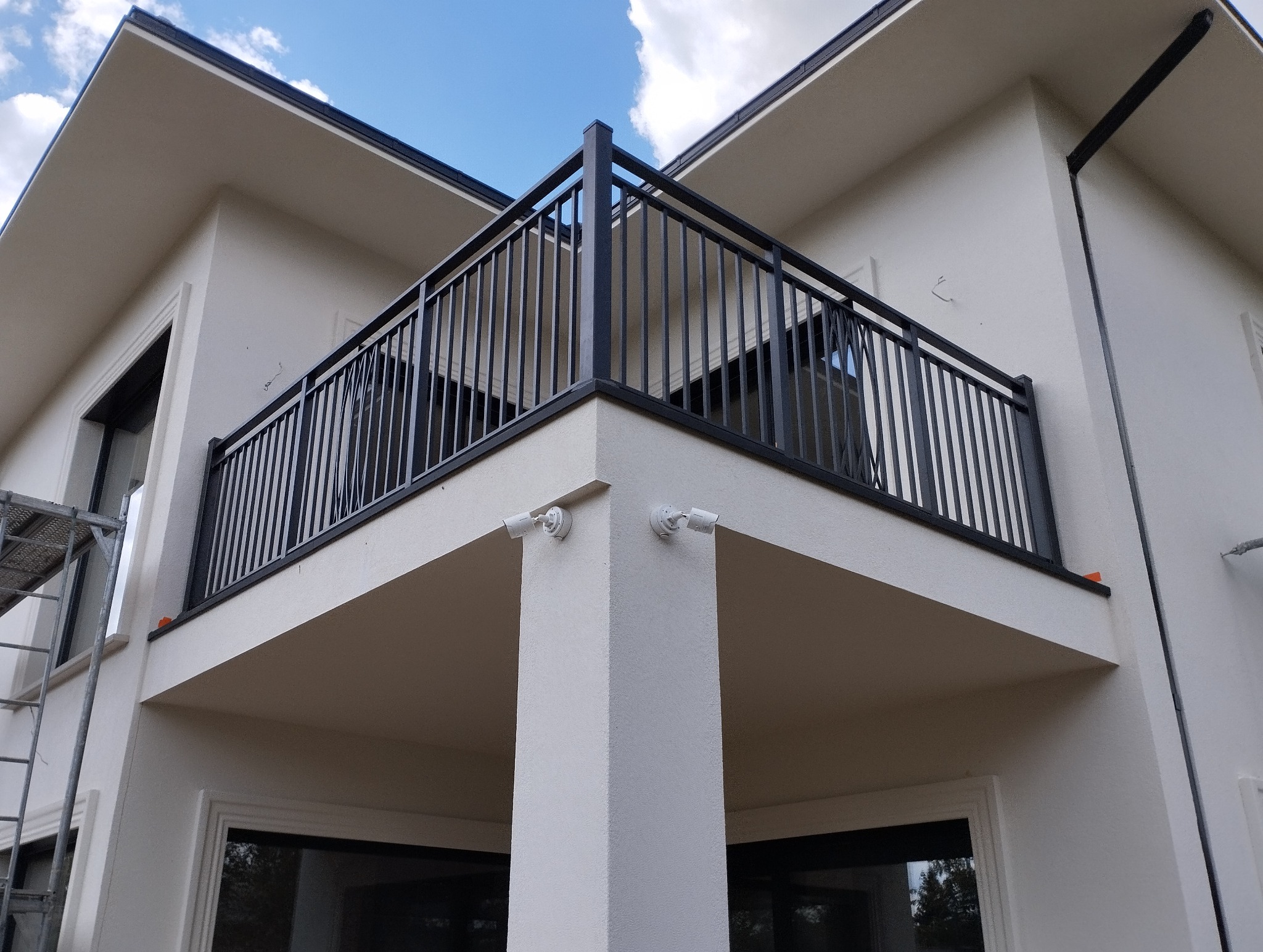 Zewnętrzny balkon z czarną metalową balustradą na tle jasnej elewacji budynku z dwoma kamerami bezpieczeństwa. Widok z dołu, częściowo widoczne niebieskie niebo.