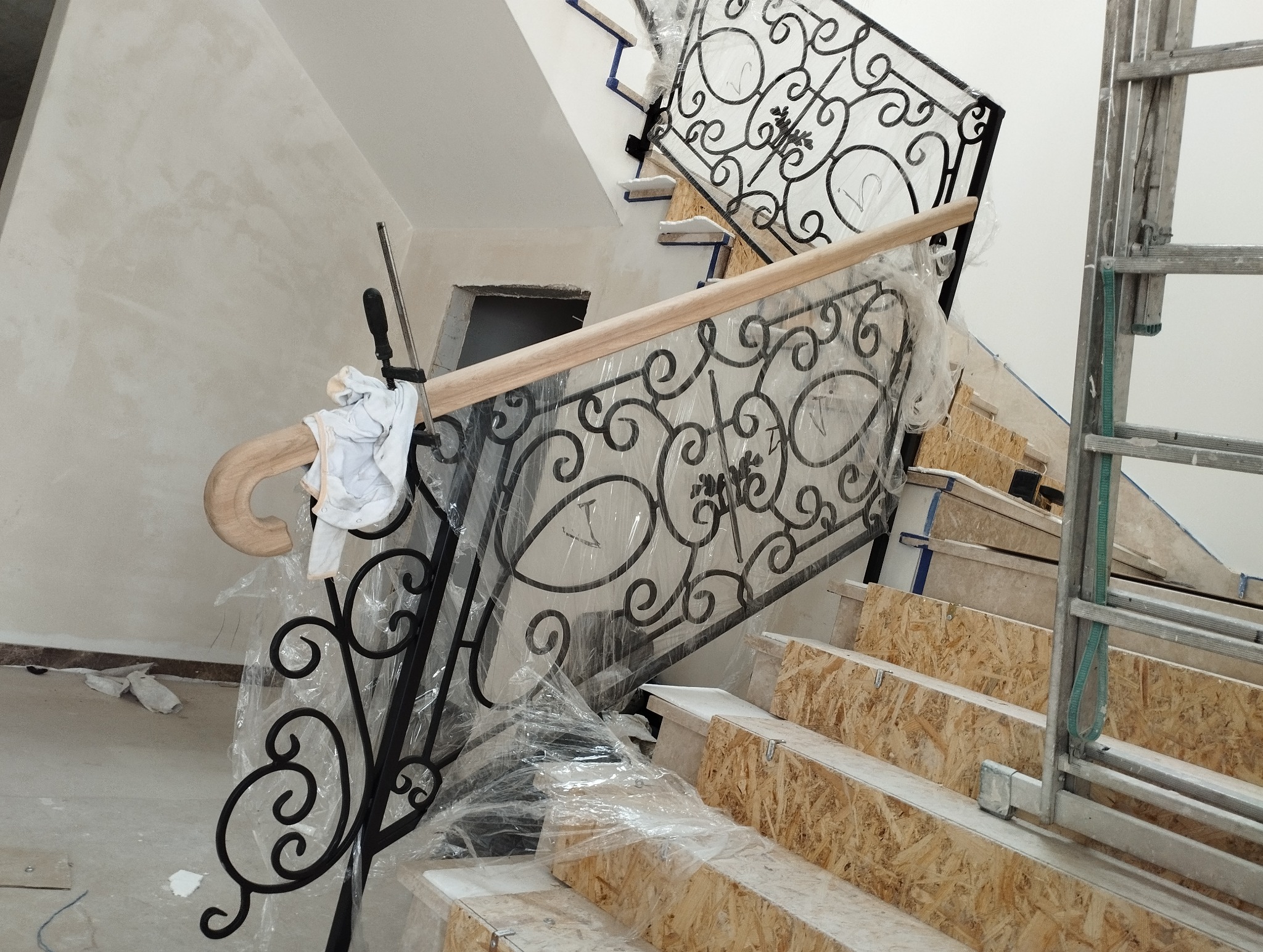 Wnętrze w trakcie prac: metalowa balustrada schodowa z drewnianym poręczą, zabezpieczona folią. Widoczne surowe stopnie schodów i drabina. Detal z bliska.