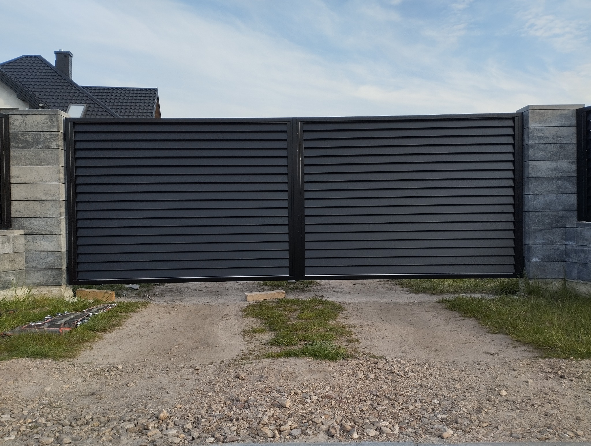 Nowoczesna, dwuskrzydłowa brama wjazdowa z poziomych paneli, w kolorze antracytowym, zamontowana na betonowych słupkach. Widoczny podjazd i fragment domu.