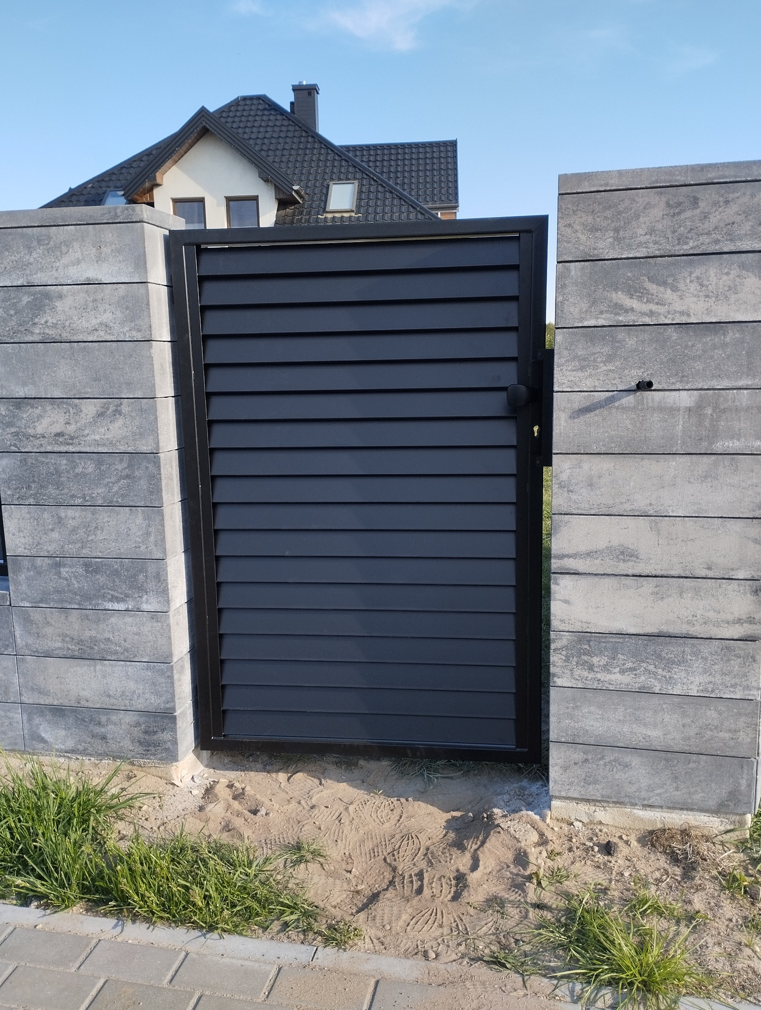 Nowoczesna, czarna furtka z poziomych profili, osadzona między dwoma słupkami z szarej, betonowej kostki. Tło: dom jednorodzinny i błękitne niebo.