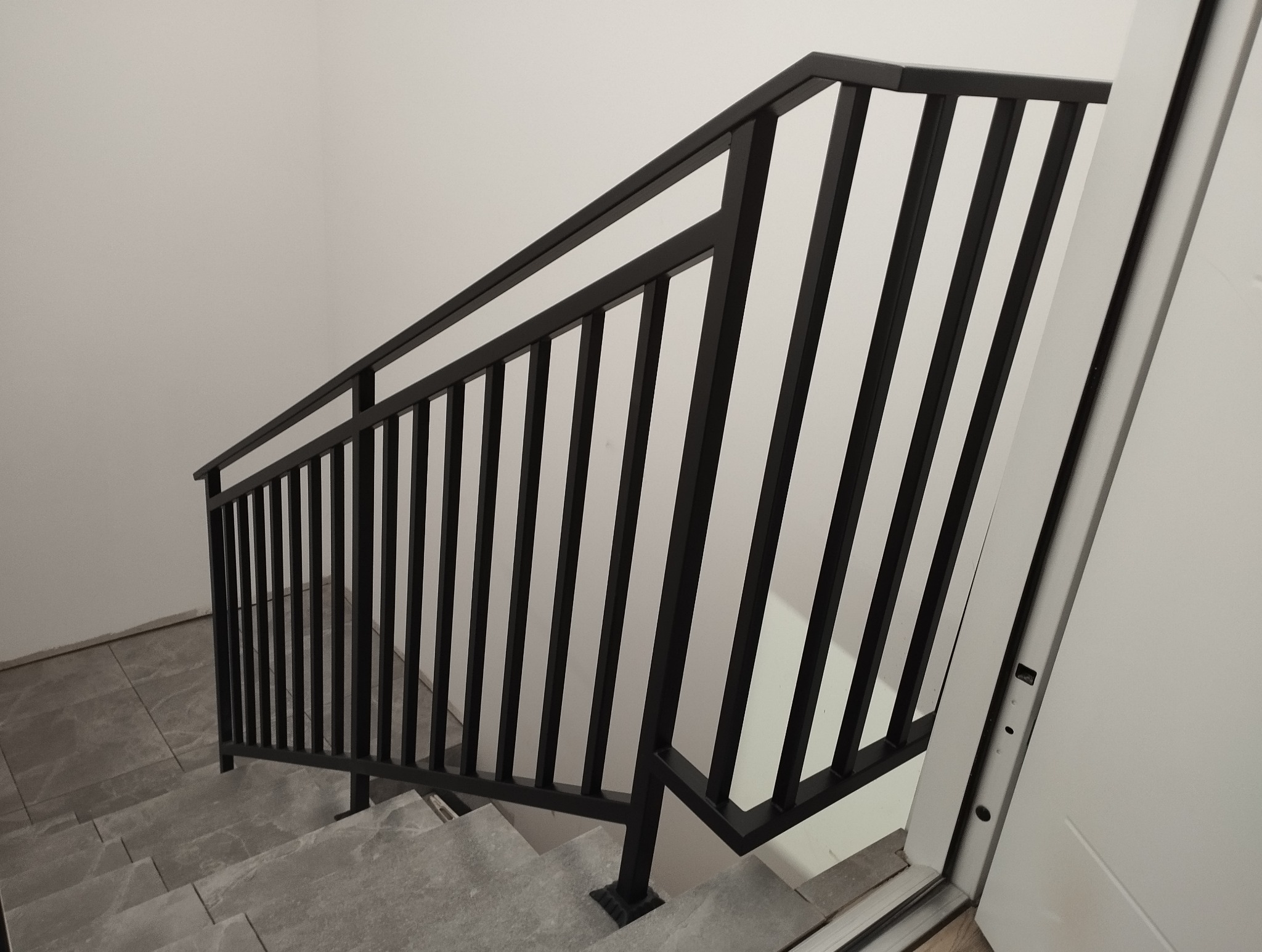 Nowoczesne schody z czarną, metalową balustradą i szarymi stopniami z płytek. Minimalistyczny design, jasne wnętrze z białą ścianą i drzwiami.