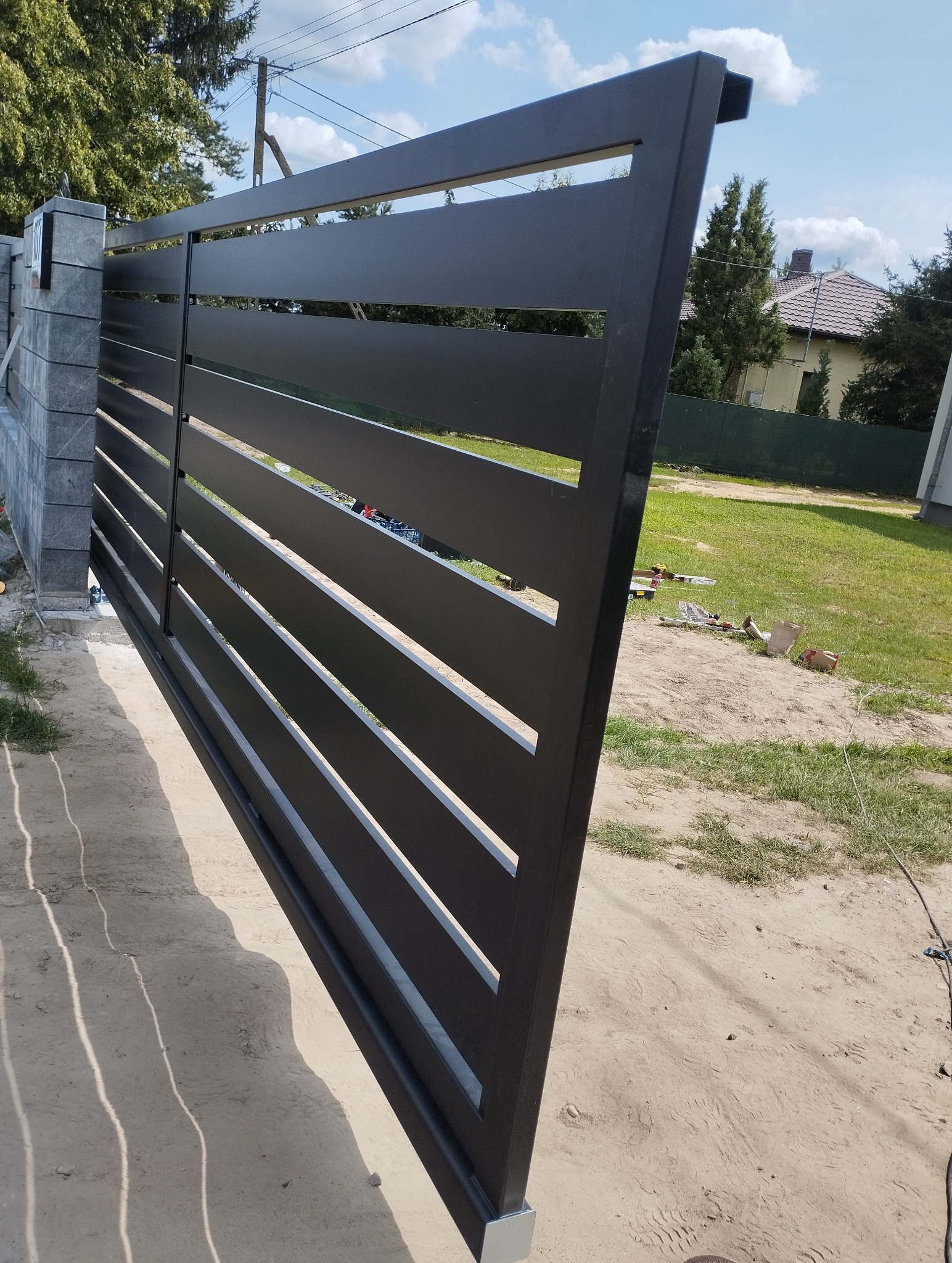 Nowoczesna, przesuwna brama wjazdowa z poziomych, czarnych profili metalowych. Widoczne narzędzia i materiały budowlane w tle sugerują etap montażu.