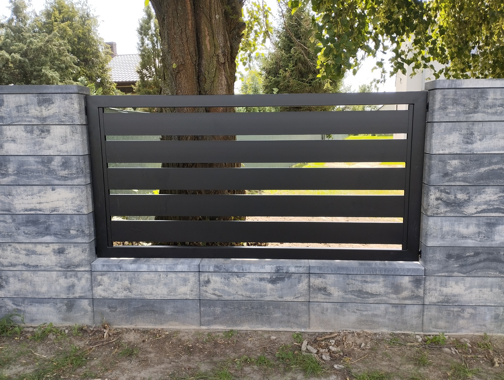 Nowoczesne, poziome przęsło ogrodzeniowe z czarnych profili, osadzone na słupkach z szarej, betonowej imitacji kamienia. W tle zieleń drzew.