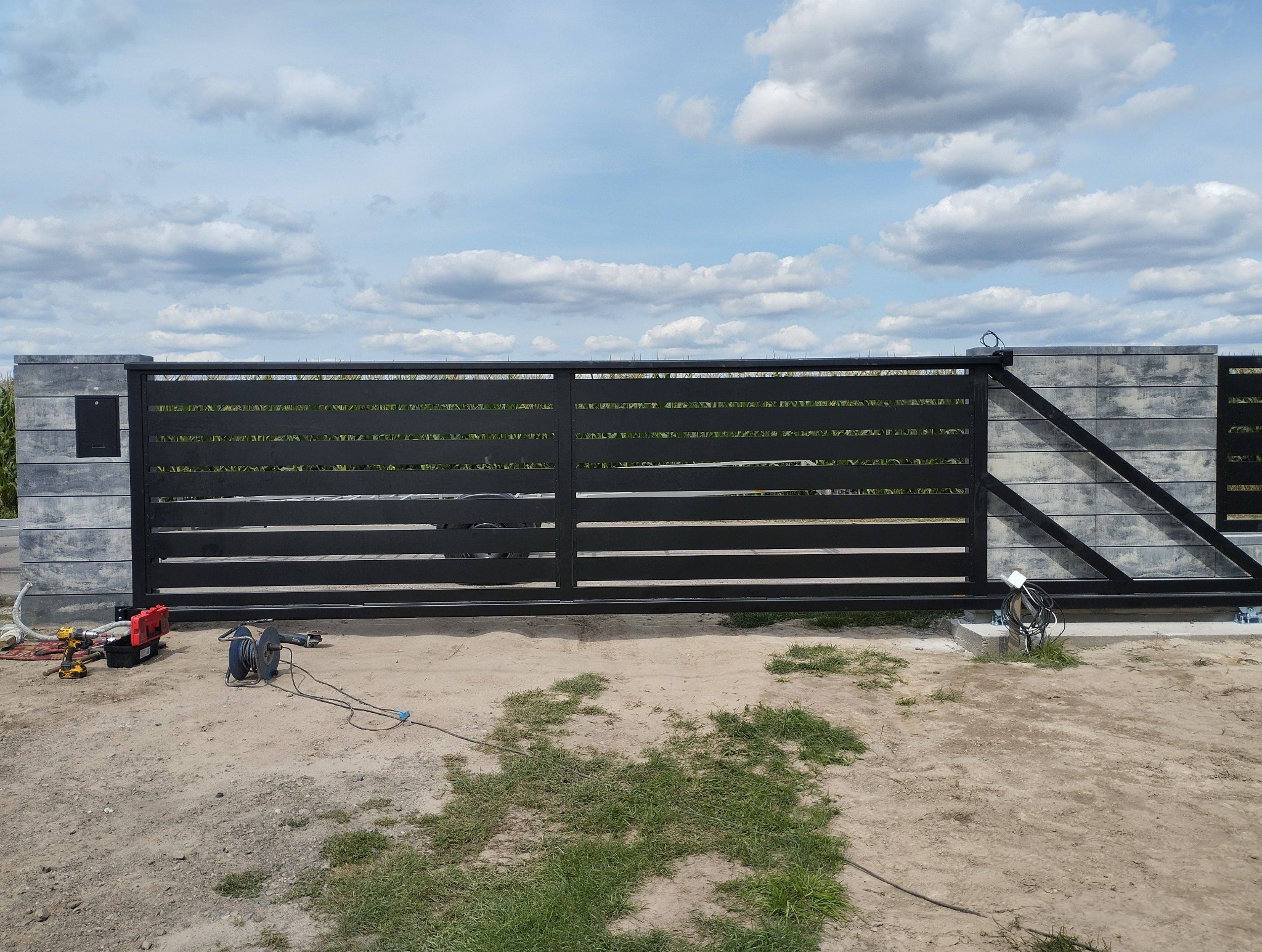 Nowoczesna, przesuwna brama wjazdowa z poziomych, czarnych profili, osadzona na betonowych słupkach. Widoczne narzędzia montażowe na ziemi. Błękitne niebo z chmurami w tle.