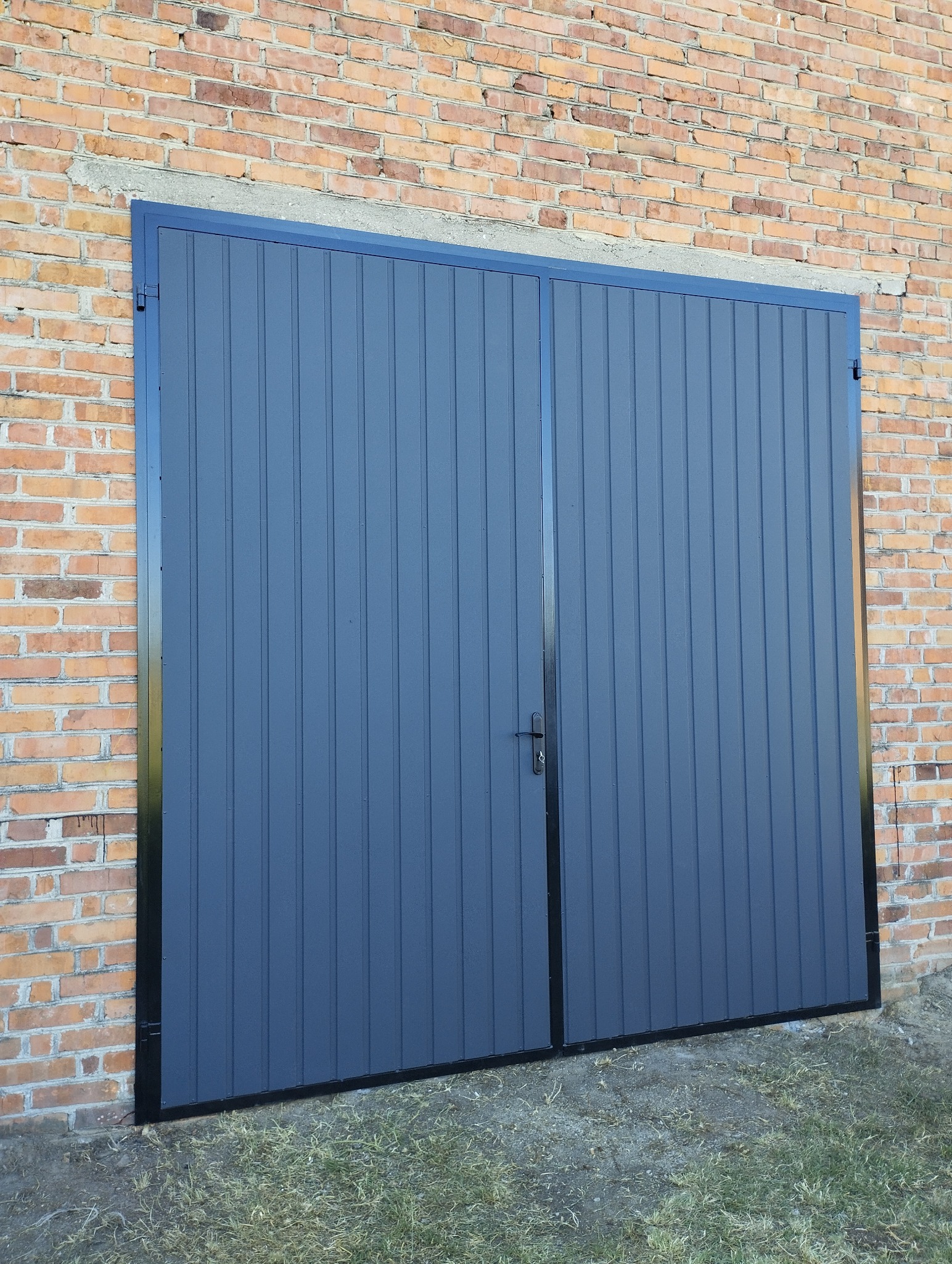 Szara, dwuskrzydłowa brama garażowa z pionowym przetłoczeniem, zamontowana w ceglanym budynku w Parczewie. Czarna rama i klamka.