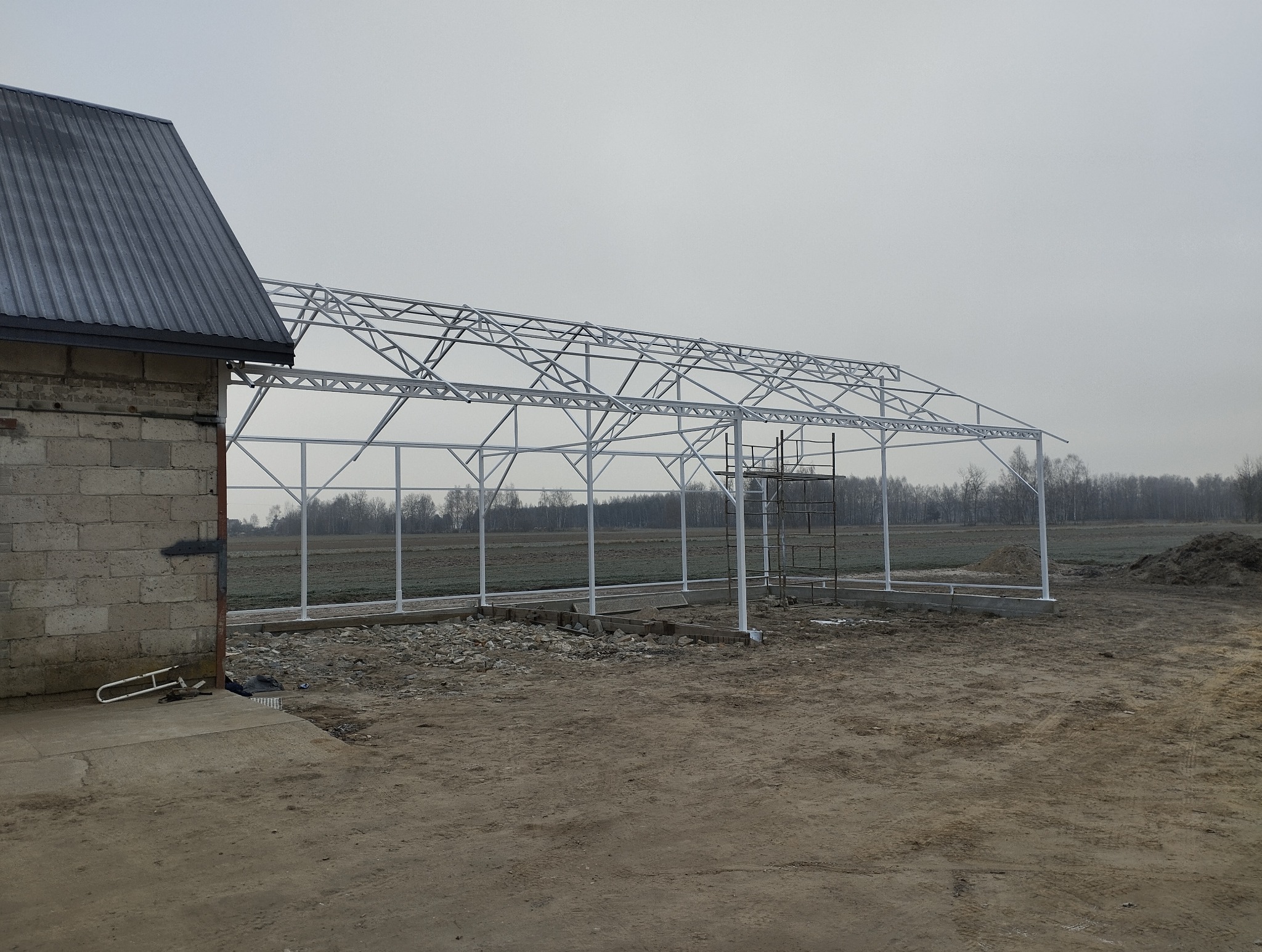 Konstrukcja metalowa szkieletu szklarni w trakcie budowy, widok z zewnątrz, obok budynek gospodarczy, pochmurny dzień.