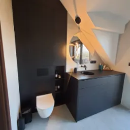 Nowoczesna łazienka na poddaszu z czarnymi meblami: szafa w zabudowie, umywalka z szafką, okrągłe lustro z oświetleniem LED. Biała toaleta i szara podłoga.