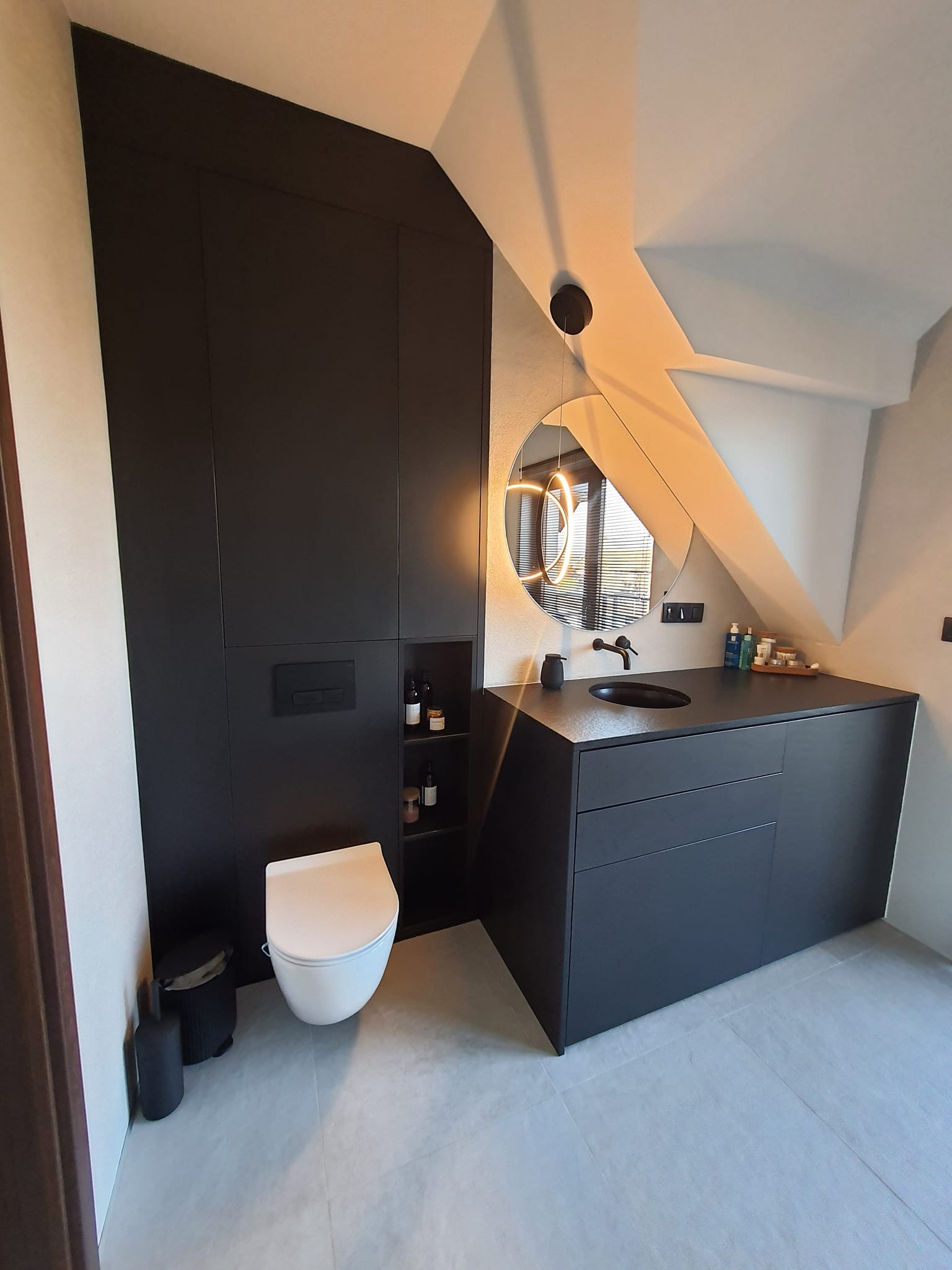 Nowoczesna łazienka na poddaszu z czarnymi meblami: szafa w zabudowie, umywalka z szafką, okrągłe lustro z oświetleniem LED. Biała toaleta i szara podłoga.