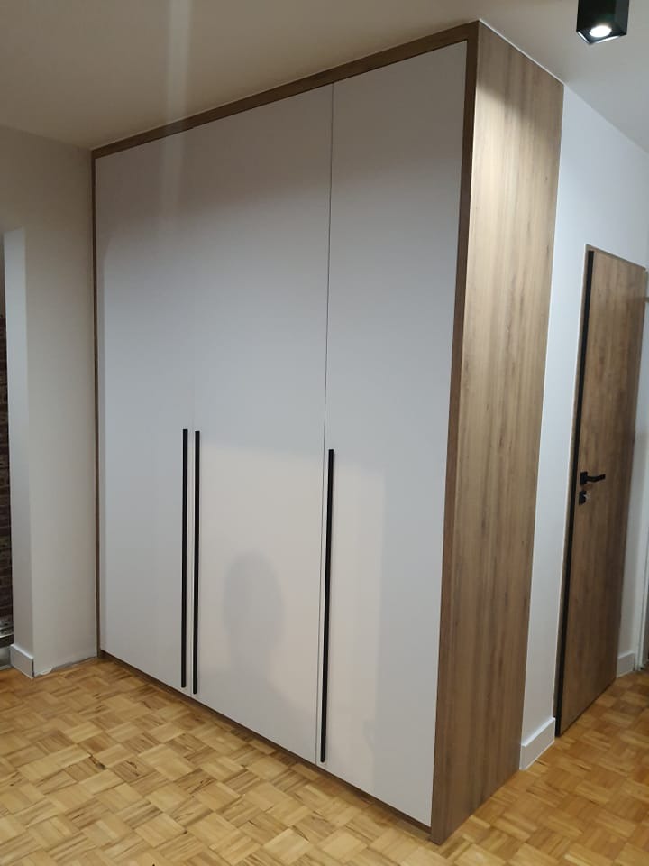 Biała szafa wnękowa z drewnianymi bokami i długimi, czarnymi uchwytami na tle parkietu.