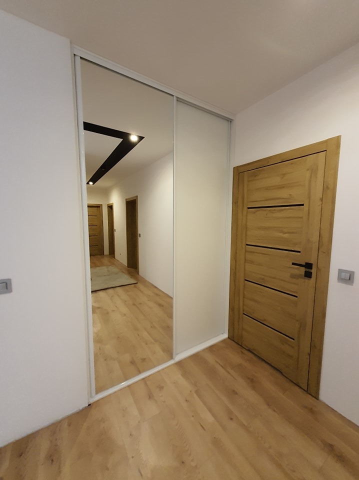Wnętrze przedpokoju z szafą wnękową z przesuwnymi drzwiami lustrzanymi i drewnianą podłogą, widoczne drewniane drzwi z czarną klamką oraz odbicie dalszej części korytarza.