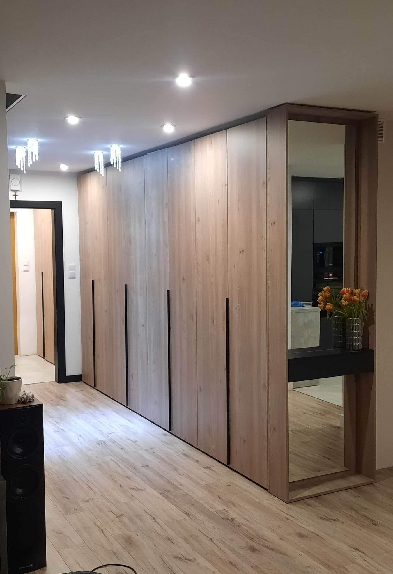 Długa szafa wnękowa z drewnianymi frontami i czarnymi uchwytami, zakończona lustrem z półką na kwiaty, widok z perspektywy podłogi.