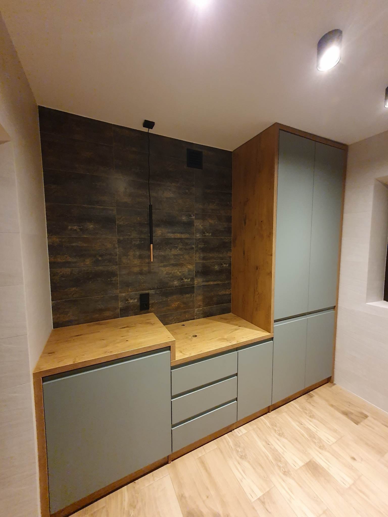 Szara zabudowa meblowa z drewnianym blatem i szafkami, na tle ściany z płytek imitujących drewno, oświetlona wiszącą lampą z elementami miedzianymi.