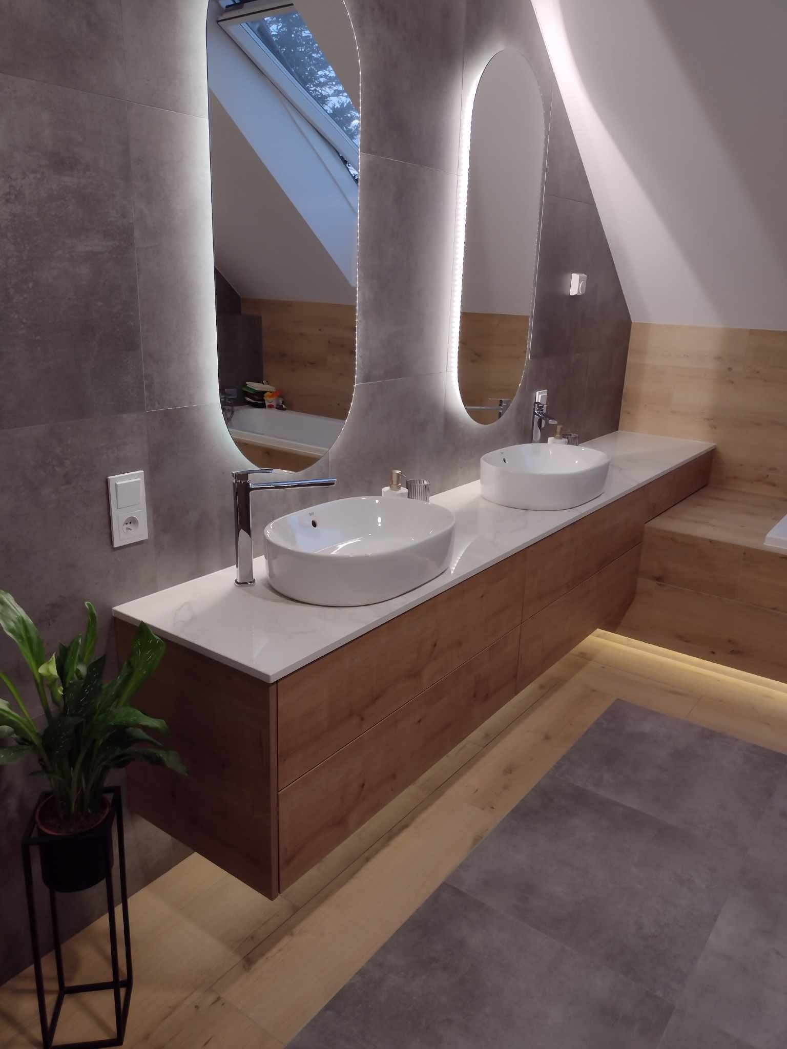 Nowoczesna łazienka na poddaszu z dwoma owalnymi umywalkami nablatowymi, podświetlanymi lustrami, drewnianą szafką pod umywalką i szarą mozaiką na ścianie.