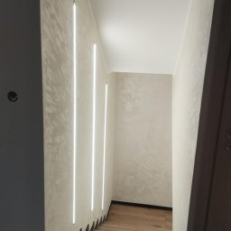 BiG-Max - Korytarz ze schodami wykończony w jasnych barwach, z dekoracyjną strukturą na ścianach i pionowymi, wbudowanymi lampami LED. Drewniane schody.