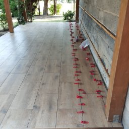 BiG-Max - Układanie płytek imitujących drewno na zewnątrz budynku, widoczne czerwone klipsy poziomujące, tło z zielenią i elementami architektury ogrodowej.