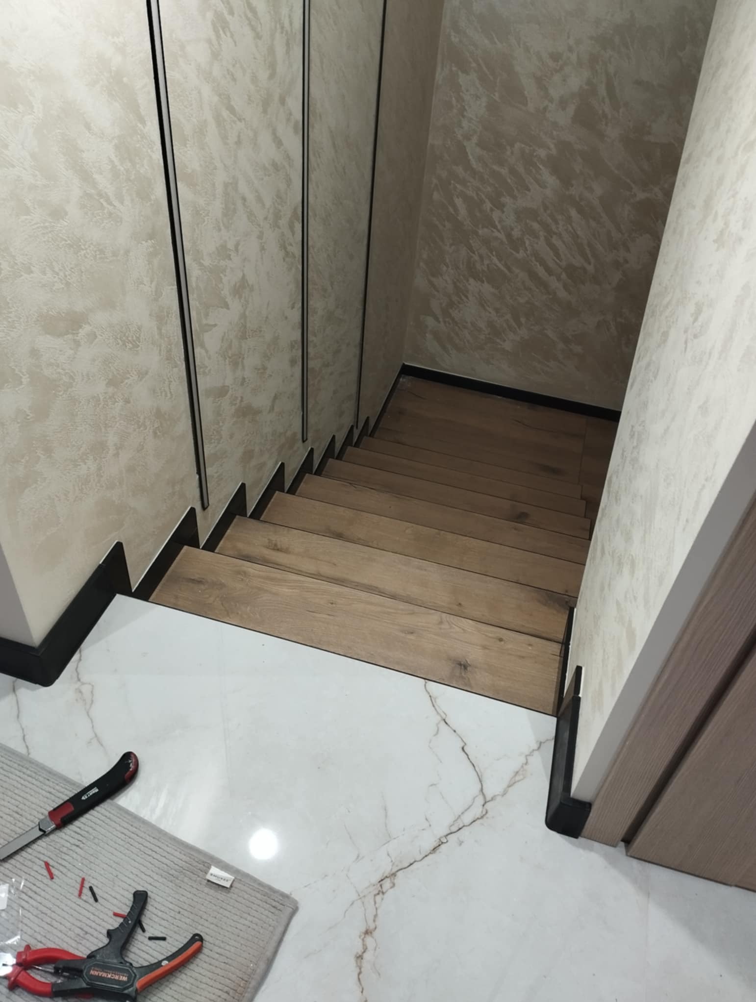 Wykończone schody z drewnopodobnymi panelami, białe marmurowe płytki na podłodze, narzędzia na szmatce. Ściany z dekoracyjnym tynkiem.