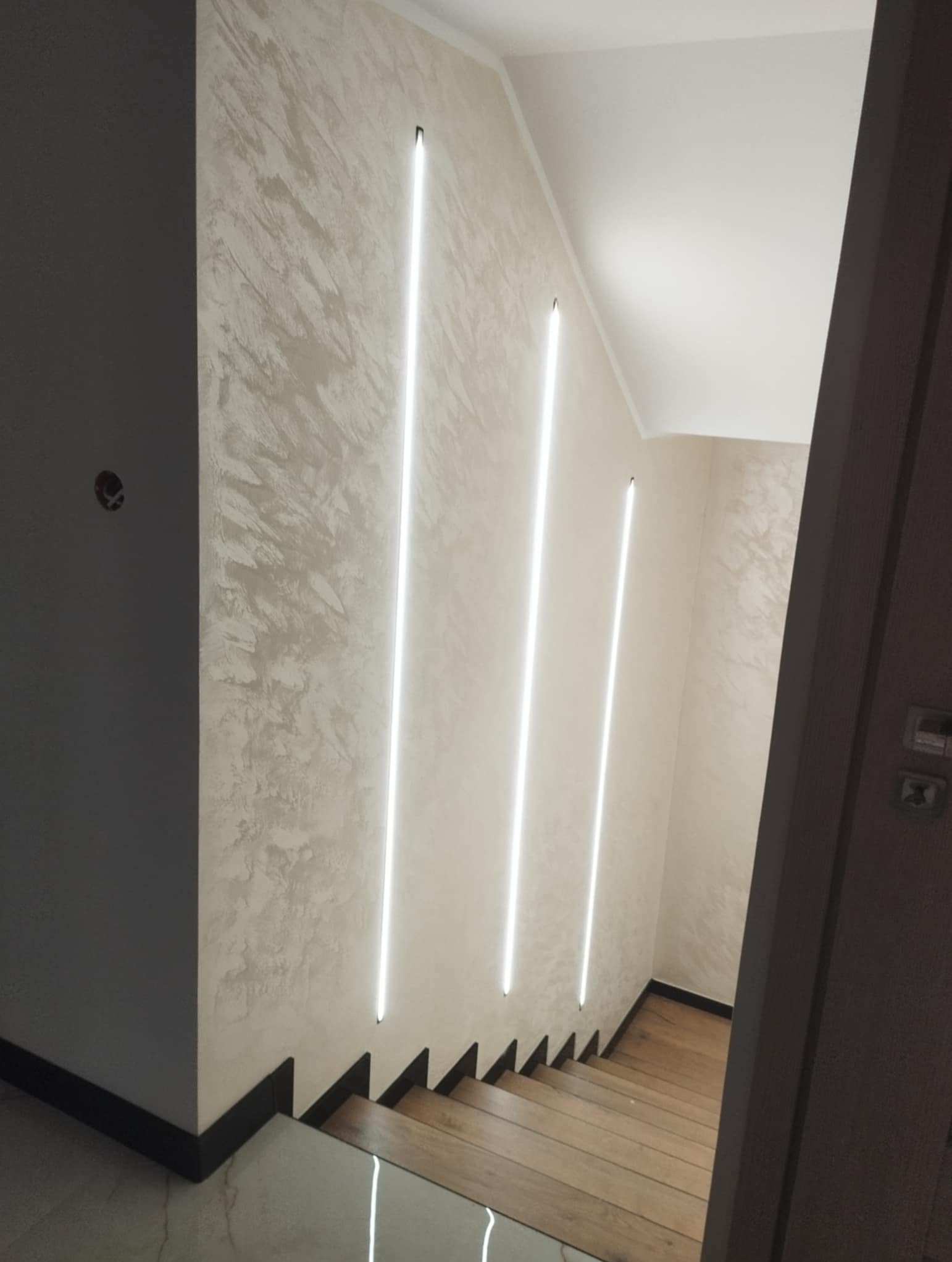 Klatka schodowa z drewnianymi schodami i dekoracyjną ścianą z oświetleniem LED. Nowoczesne wykończenie wnętrza z akcentem na pionowe linie świetlne.