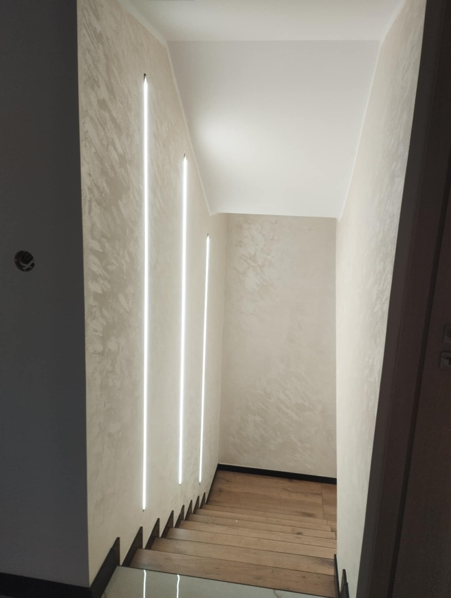 Korytarz ze schodami wykończony w jasnych barwach, z dekoracyjną strukturą na ścianach i pionowymi, wbudowanymi lampami LED. Drewniane schody.